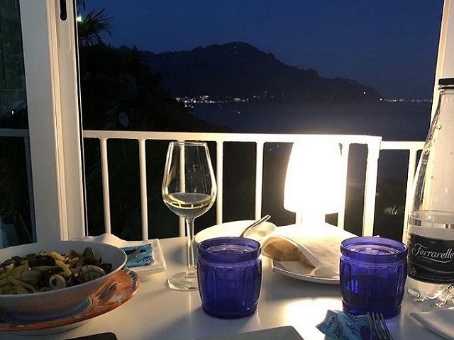 Tavolo serale in terrazza, con un calice di vino bianco e le luci della costa